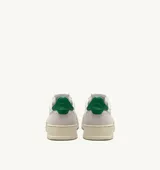 Championes de estilo vintage inspirados en los años 80, confeccionados en cuero blanco con paneles de gamuza gris en la puntera y el talón. Presentan perforaciones en la puntera para mayor ventilación, cordones planos de algodón y un detalle en color verde en el talón. La suela es de goma en tono crema con el logo de la marca.
