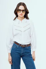 Camisa blanca de manga larga abullonada con puños elásticos, cuello camisero clásico y cierre frontal de botones. Presenta detalles de alforzas y encaje geométrico en la pechera.