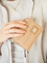 Monedero de lona color beige con cierre perimetral, pespuntes decorativos verticales y parche frontal con logo de la marca.