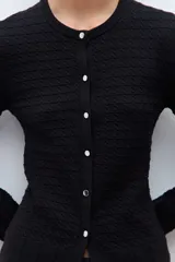 Cardigan negro de manga larga con cuello redondo y cierre frontal de botones. Presenta un tejido con patrones estructurados y un borde festoneado en el dobladillo.