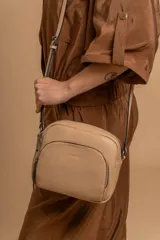 Bandolera de diseño compacto en color beige, confeccionada en material símil cuero con textura granulada. Cuenta con un bolsillo frontal con cierre y correa larga regulable.