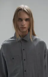 Camisa de manga larga en tejido twill color gris melange, con corte oversize. Presenta cuello clásico, cierre frontal con botones y tres bolsillos delanteros, uno de ellos oculto.