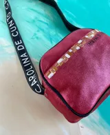 Bandolera Kaira de la línea Ottenta de Carolina De Cunto, en color fucsia metalizado. Cuenta con un diseño compacto, bolsillo exterior con broche imantado y detalle pintado a mano, cierre superior y una correa larga regulable con el logo de la marca estampado.