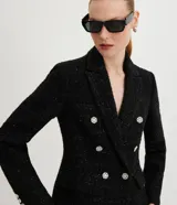 Blazer negro de tweed con hilos metalizados, corte entallado, escote en V profundo, bolsillos delanteros con solapa y doble botonadura con seis botones decorativos de strass.