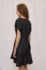 Vestido corto de algodón color blanco, con escote en V y botonadura frontal. Presenta mangas cortas holgadas, cintura ajustada con frunces y falda acampanada con terminación de volado en el ruedo.