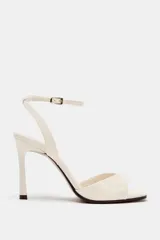 Sandalia de tacón alto en color blanco marfil, con tira delantera ancha, diseño destalonado y tira ajustable al tobillo con hebilla metálica dorada. Presenta punta cuadrada y tacón fino.