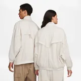 Chaqueta Nike Sportwear Essential Windrunner de color beige, diseño oversize con cierre frontal completo y cuello alto. Confeccionada en tejido arrugado ligero con forro de malla, presenta el logotipo de la marca bordado en el pecho.