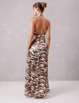 Vestido largo de tejido satinado plisado con estampado animal print tipo cebra en tonos tierra y ocre. Presenta escote halter profundo en V con drapeado central y broche de pedrería, espalda descubierta y falda fluida con apertura frontal.