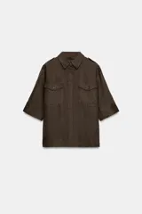 Camisa de lino color marrón oscuro con cuello solapa, manga por debajo del codo y cierre frontal con botones ocultos por solapa. Presenta bolsillos de plastrón con solapa en el pecho y trabillas en los hombros.