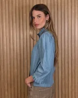 Camisa de algodón color blanco con rayas verticales rosadas, cuello alto con volados, mangas tres cuartos abullonadas con puños elastizados con volados y cierre frontal de botones oscuros.