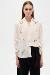Camisa de algodón blanca de corte holgado, con cuello camisero, mangas largas y bordados florales multicolores. Presenta detalles de calados en los hombros y cierre frontal con botones.