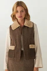 Chaleco corto de corte recto con diseño de pata de gallo en tonos marrones. Presenta cuello y detalles en los bolsillos de piel sintética, con aplicaciones de cuero en la parte inferior de los bolsillos.