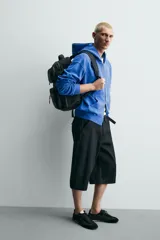 Mochila técnica de color negro con múltiples compartimentos, incluyendo dos bolsillos frontales con cierre, bolsillos laterales y compartimento principal acolchado para dispositivos electrónicos. Cuenta con parte posterior acolchada, banda de sujeción para equipaje, asa de mano y asas de hombro ajustables.