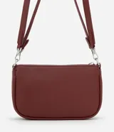 Cartera tipo baguette color bordó, confeccionada en poliuretano con acabado liso. Presenta una cadena decorativa plateada en el frente, cierre superior con cremallera y correa larga ajustable y desmontable.