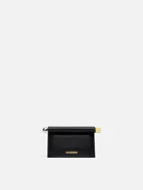 Bolso de cuero color negro modelo La Petite Pochette Rond C de Jacquemus, con silueta de sobre, solapa superior enrollada con broche magnético, extremos metálicos en plata y oro, y logo grabado en letras doradas en el frente.