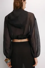 Cazadora semitransparente de color negro, con capucha, cierre frontal de cremallera metálica y mangas largas abullonadas con puños elásticos. Presenta un diseño holgado con bajo elástico.