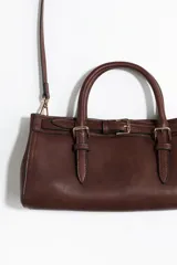 Cartera bandolera rectangular de color marrón, con diseño alargado y asas cortas de mano. Incluye un detalle de tira decorativa con hebillas metálicas en la parte frontal y herrajes dorados.