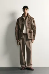 Pantalón relaxed fit de hombre color marrón con textura estructurada, cintura con pliegues frontales, bolsillos laterales y cierre frontal con cremallera y botón.