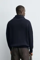 Chaqueta de punto con textura acanalada, cuello subido y cierre frontal completo con cremallera metálica. Presenta un corte relajado y acabados en rib en puños y ruedo.
