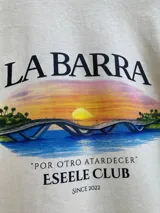 Remera oversize de color arena, 100% algodón, con estampa en la espalda que ilustra el puente de La Barra durante el atardecer, acompañada de texto descriptivo y de la marca Eseele Club.