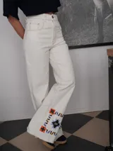 Pantalón vaquero de corte recto y tiro alto en color blanco marfil, con detalles de costuras en contraste y estampa decorativa en la parte inferior de las piernas, que incluye figuras de caballos y patrones geométricos. Presenta aberturas laterales en los tobillos.
