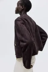 Chaqueta corta de gamuza sintética en color marrón oscuro, con forro interior de peluche. Presenta cuello redondo, cierre frontal con botones metálicos dorados y dos bolsillos delanteros con solapa y botón.