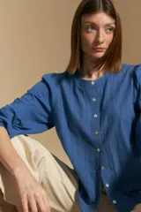 Camisa de algodón blanca con delicadas líneas bordadas verticales y textura calada. Presenta escote redondo con cierre frontal de botones, mangas largas con leve volumen en los hombros y puños elásticos.