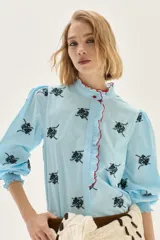 Blusa de algodón en color celeste con estampado floral en negro. Presenta cuello alto con volados y borde en color rojo, mangas abullonadas con puños elásticos tipo smock y cierre frontal con botones.