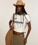 Remera estilo ringer de algodón color blanco crudo, con cuello redondo y ribetes en contraste azul marino en el cuello y mangas. Presenta un estampado frontal con la palabra "Santorini" y un gráfico estilizado.