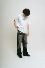 Pantalón de jean negro con lavado localizado (efecto tie-dye o acid wash), corte baggy y amplio. Presenta apliques bordados en la parte trasera de los bolsillos y herrajes metálicos cromados.