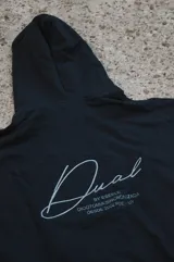 Canguro negro con capucha, corte oversize y diseño con hombros caídos. Presenta detalles bordados en la espalda con la palabra "Dual" en tipografía cursiva junto a texto descriptivo en tonos monocromáticos.