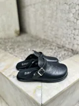 Zueco negro de cuero sintético con textura, estilo cerrado y tira ajustable con hebilla metálica plateada.