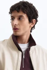 Campera bomber en mezcla de algodón y nylon color beige, con cuello elevado acanalado y cierre frontal de cremallera. Cuenta con bolsillos laterales y un bolsillo con cierre en la manga izquierda. Puños y bajo en punto acanalado, de corte holgado.