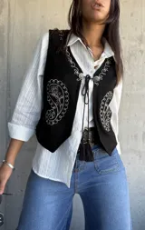 Chaleco negro de gamuza con bordados blancos estilo paisley y cierre frontal con cordones y detalles metálicos.