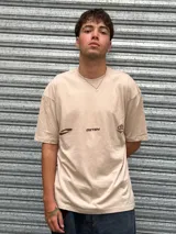 Remera de corte oversize color beige, con cuello redondo y mangas cortas. Presenta un diseño gráfico estampado en el pecho con la palabra 'OSTEN' y logotipos abstractos en color marrón.