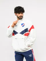 Buzo deportivo de nylon con medio cierre frontal y cuello alto. Presenta un diseño de bloques de color en azul, blanco y bordó, con el escudo del Club Nacional de Football y el logo de Umbro en el pecho.
