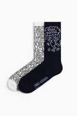 Pack de dos pares de medias de caña alta con diseño artístico inspirado en Keith Haring. Un par es blanco con estampado integral de figuras y el otro es negro con un gráfico central que dice 'Rain Dance'.