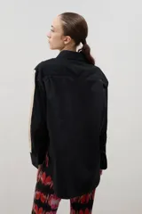 Camisa de corte holgado y largo medio, color negro con detalle de franja lateral en color crema a lo largo de las mangas. Presenta cuello clásico, cierre frontal con botones y tajos laterales en el ruedo.