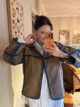 Chaqueta de paño de lana color marrón con detalles de costuras artesanales en crochet en color azul y verde. Presenta un diseño de corte recto y holgado con solapas.