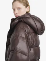 Campera puffer de corte holgado en color marrón oscuro con acabado brillante. Presenta cuello alto, cierre frontal con cremallera y botones a presión, además de bolsillos frontales con solapa.