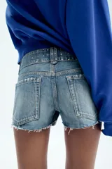 Shorts de denim de tiro bajo con cinco bolsillos, detalle de tachas, aplicación de brillos, bajo deshilachado y cierre frontal con cremallera y botones metálicos.