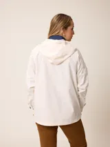 Campera de corte recto y estilo funcional, color beige, con capucha integrada, cierre frontal con botones a presión y bolsillos delanteros amplios. Presenta un diseño minimalista con cordones ajustables en la capucha.
