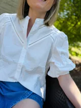 Camisa blanca de popelina de algodón, manga corta abullonada y cuello mao. Presenta doble detalle de puntilla en forma de V en la parte superior delantera.