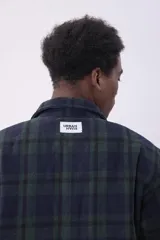 Campera tipo sobrecamisa de franela con diseño a cuadros en tonos beige y marrón. Presenta cuello clásico, cierre frontal con botones, dos bolsillos frontales con solapa y botones en el pecho, y mangas largas con puños abotonados.