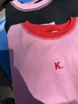 Buzo deportivo oversize color rosa, con cuello redondo y costuras expuestas en rojo. Presenta un pequeño bordado de una letra 'K' en el pecho.
