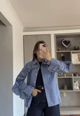 Camisa de corte recto con estampado de cuadros vichy en tonos azul y blanco. Presenta cuello clásico, cierre frontal con botones y volados decorativos que recorren la parte superior de las mangas y los hombros.