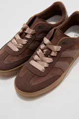 Zapatillas urbanas de corte bajo en tonos beige y tostado, combinando paneles de cuerina y gamuza sintética. Cuentan con cordones anchos tipo cinta de raso en color crudo, pespuntes decorativos y suela de goma al tono.