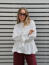 Blusa blanca de textura arrugada, con escote en V y lazos para anudar. Presenta mangas largas con puños acampanados y un diseño con volado en el ruedo.