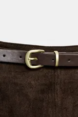Falda midi de tejido efecto ante en color marrón oscuro, con tiro medio, trabillas en la cintura y cinturón ajustable con hebilla metálica. Presenta un diseño de bajo recto con una abertura frontal central y cierre lateral con cremallera oculta.