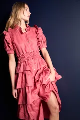 Vestido largo color rosado con volados en mangas y falda con capas. Tiene cuello camisero, mangas cortas abullonadas y cinturón de lazo en la cintura.